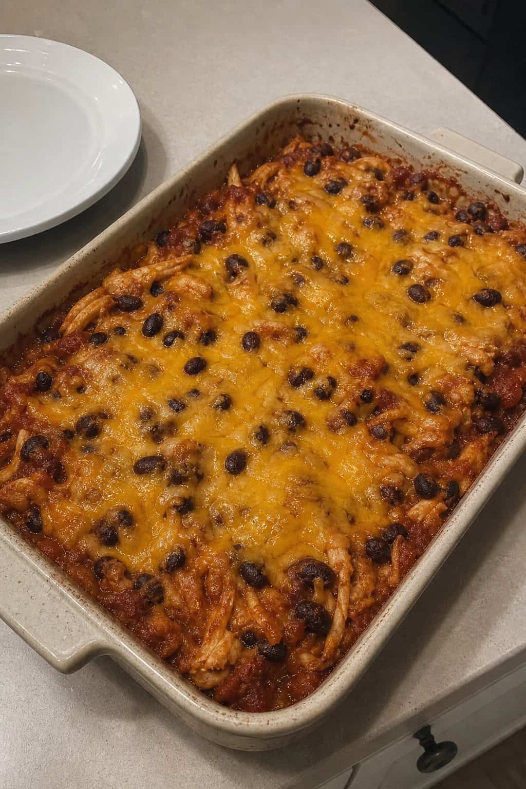 A baked enchilada casserole with ground turkey, black beans, and melted cheese in a ceramic dish, with small bowls of sliced avocado, sour cream, lime wedges, and diced onion visible on the wooden table.