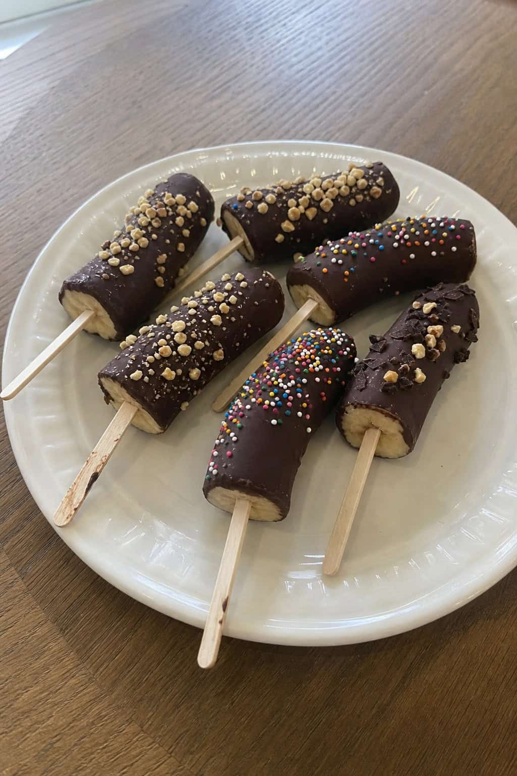 Several chocolate-dipped frozen banana pops arranged on a wooden board with banana slices and a bowl of melted chocolate nearby.