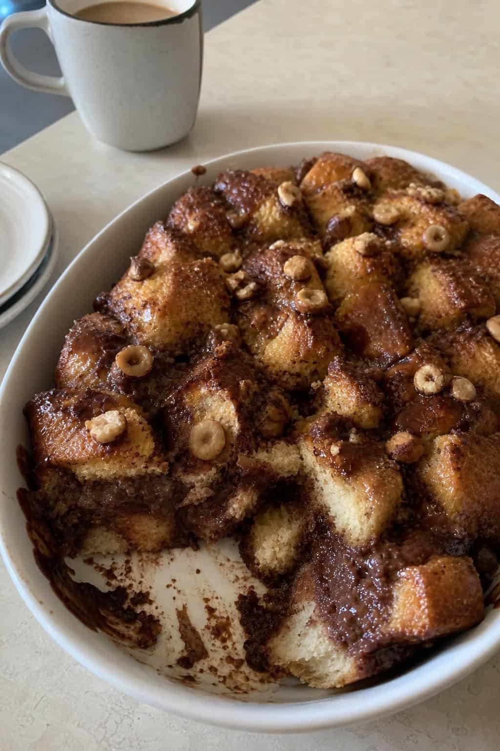 A chocolate-hazelnut French toast pull-apart casserole on a wooden table with some pieces separated to show the gooey interior, surrounded by whole hazelnuts, a jar of chocolate spread, and a coffee cup.