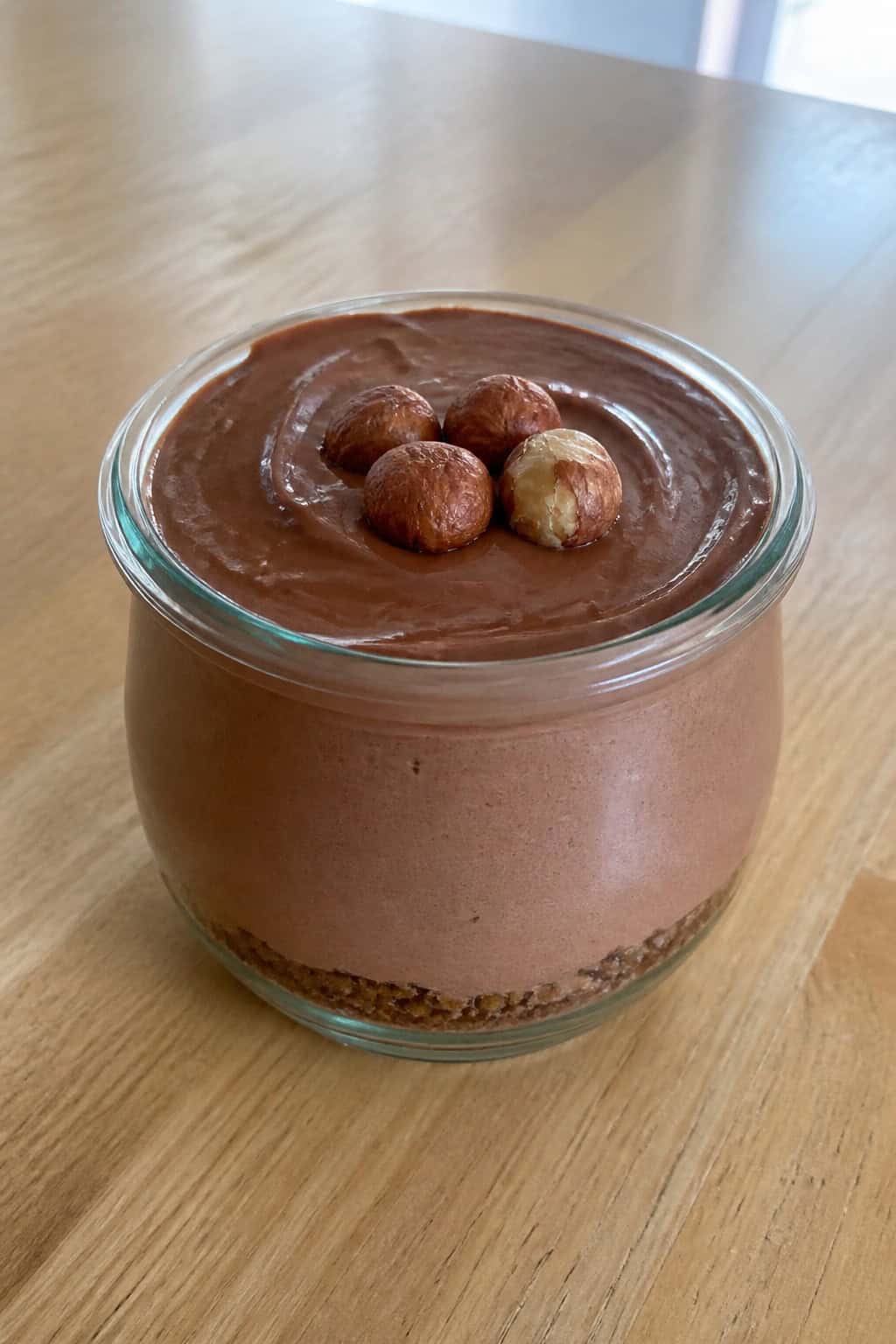 A glass jar filled with chocolate hazelnut keto cheesecake pudding topped with hazelnuts and a mint leaf on a wooden table.