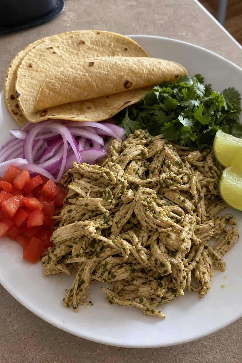 A plate of shredded cilantro lime chicken with fresh taco ingredients like tomatoes, onions, cilantro, lime wedges, and corn tortillas on a table.