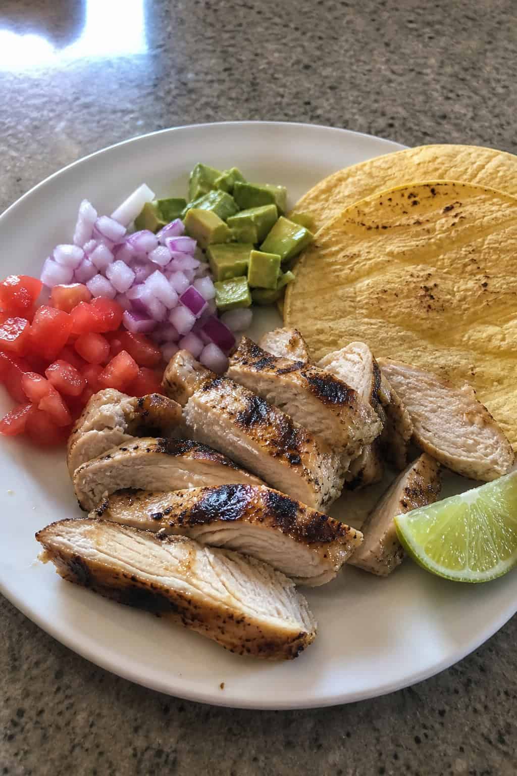 A plate of grilled chicken breast garnished with cilantro and lime wedges, surrounded by taco ingredients like diced onions, tomatoes, avocado, and tortillas.