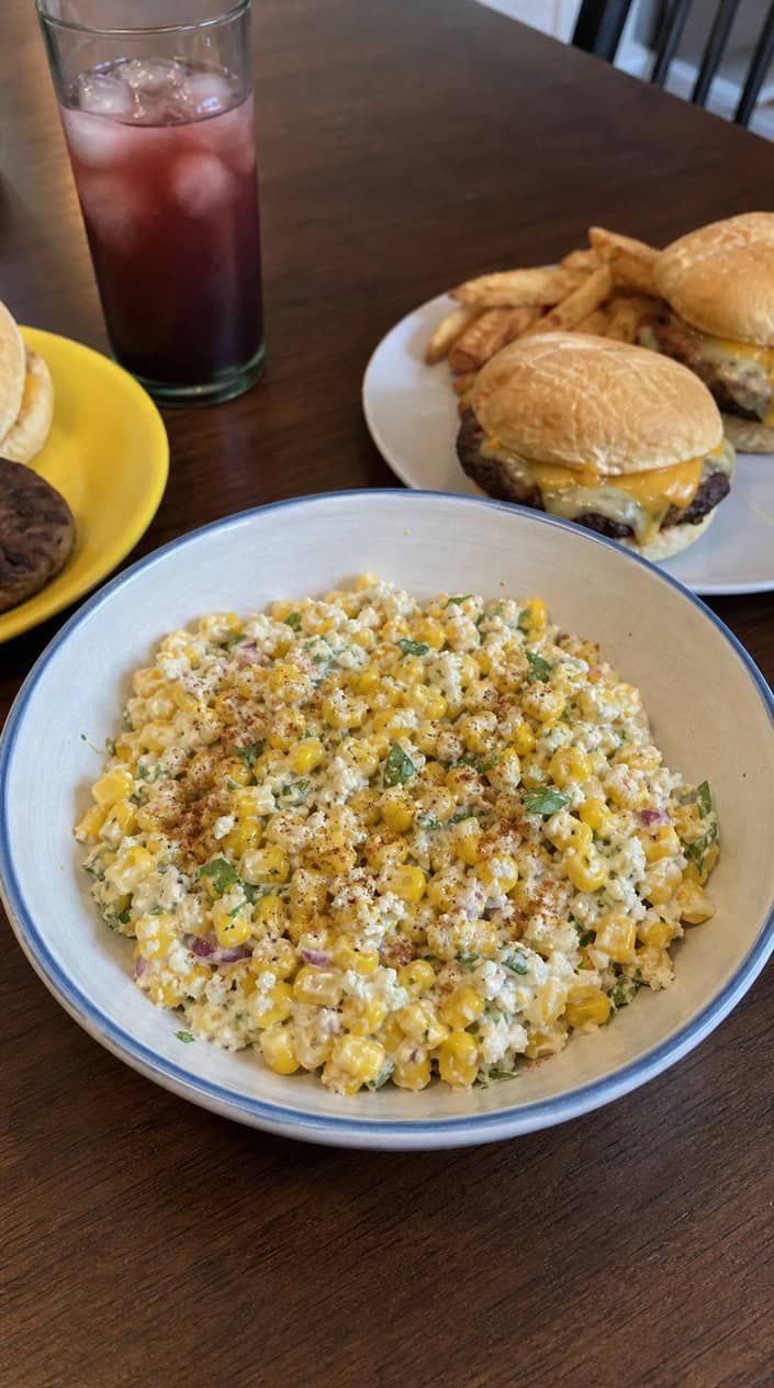 A bowl of Mexican street corn salad with corn, cilantro, and lime wedges on a wooden table.