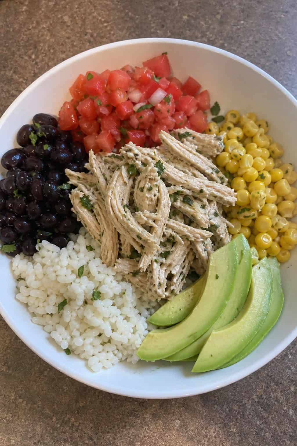 A wide bowl on a wooden table layered with white rice, black beans, corn, shredded cilantro-lime chicken, fanned avocado slices, and a spoonful of bright red pico de gallo.