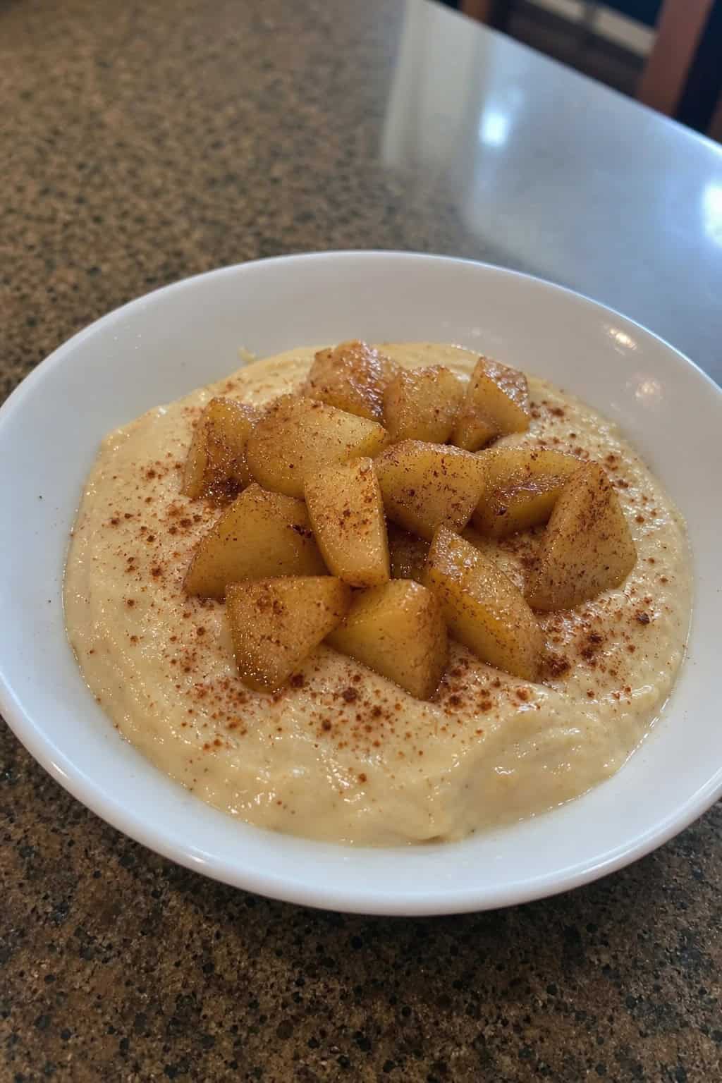 A bowl of cinnamon apple keto cheesecake pudding topped with stewed apple slices and a sprinkle of cinnamon on a wooden surface with cinnamon sticks and apples in the background.