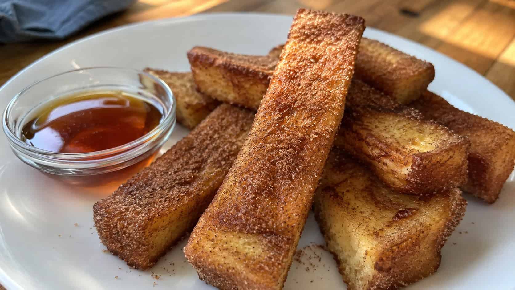 finished cinnamon French toast sticks stacked on a white plate, golden browned edges rolled in cinnamon sugar, a small bowl of maple syrup beside them for dipping, warm natural morning light, casual home cooking