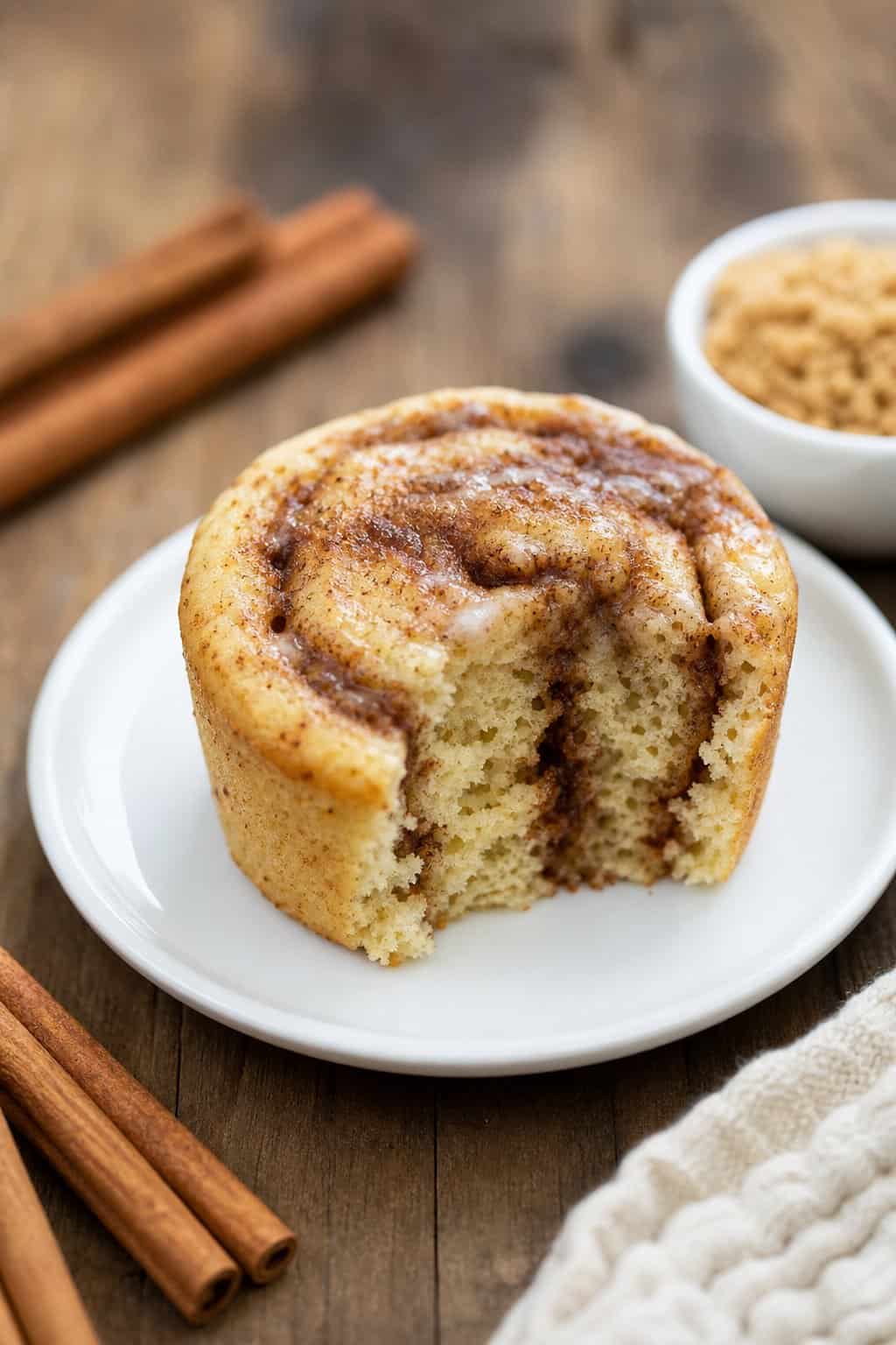 A cinnamon roll mug muffin served on a small white plate, with a visible cinnamon swirl on the top and two cinnamon sticks resting against the mug on a warm-toned wooden table.