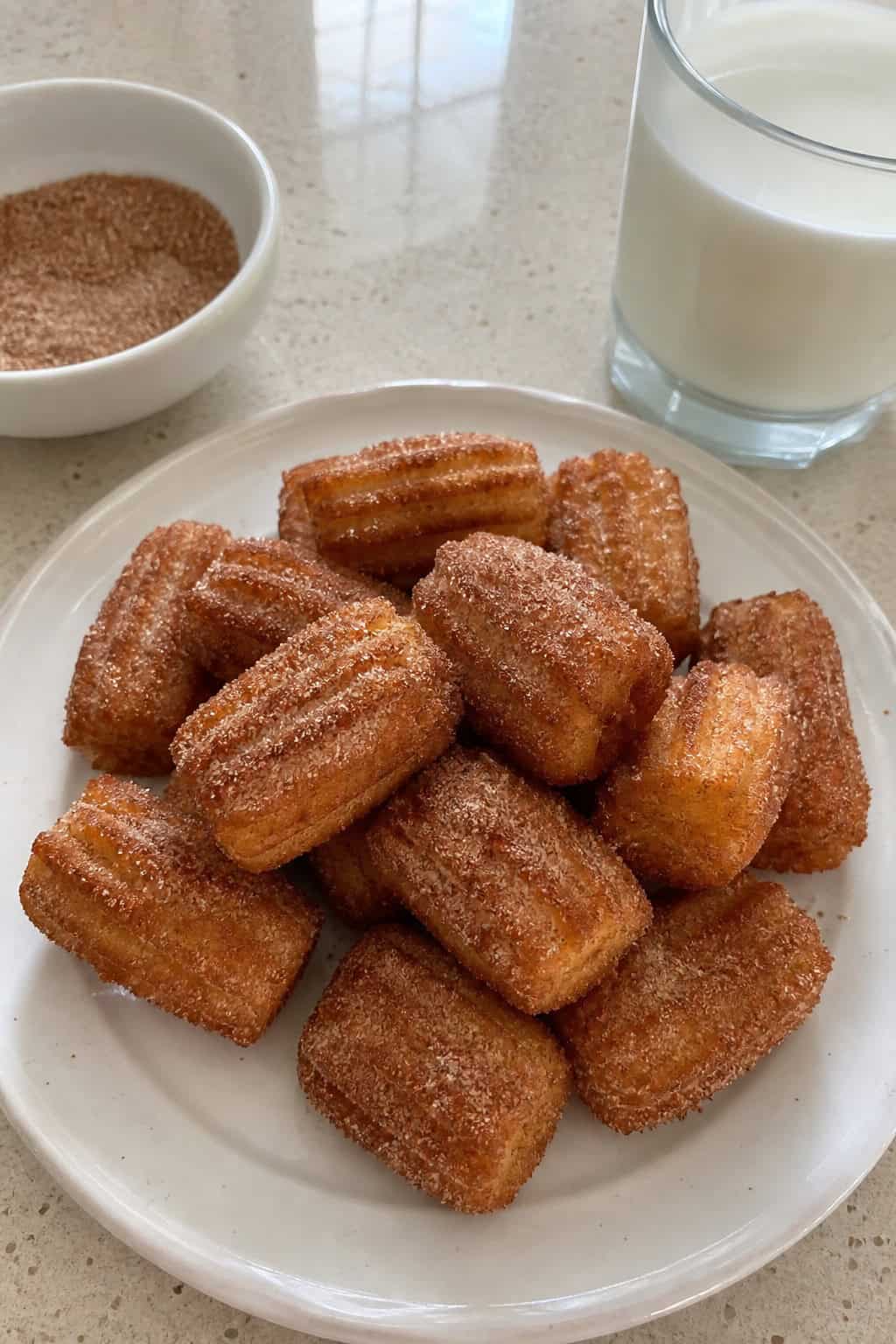 Close-up of cinnamon sugar churro bites on a plate with a bowl of cinnamon sugar and a glass of milk on a wooden table.