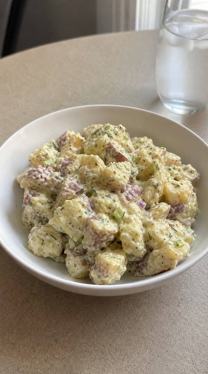 A bowl of creamy potato salad garnished with dill and mustard seeds on a wooden table with a blurred backyard barbecue scene in the background.