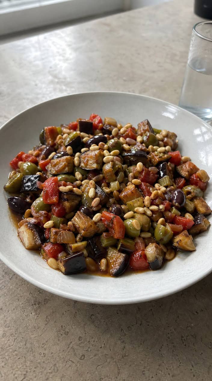 A bowl of classic caponata with eggplant and pine nuts on a wooden table surrounded by fresh ingredients.