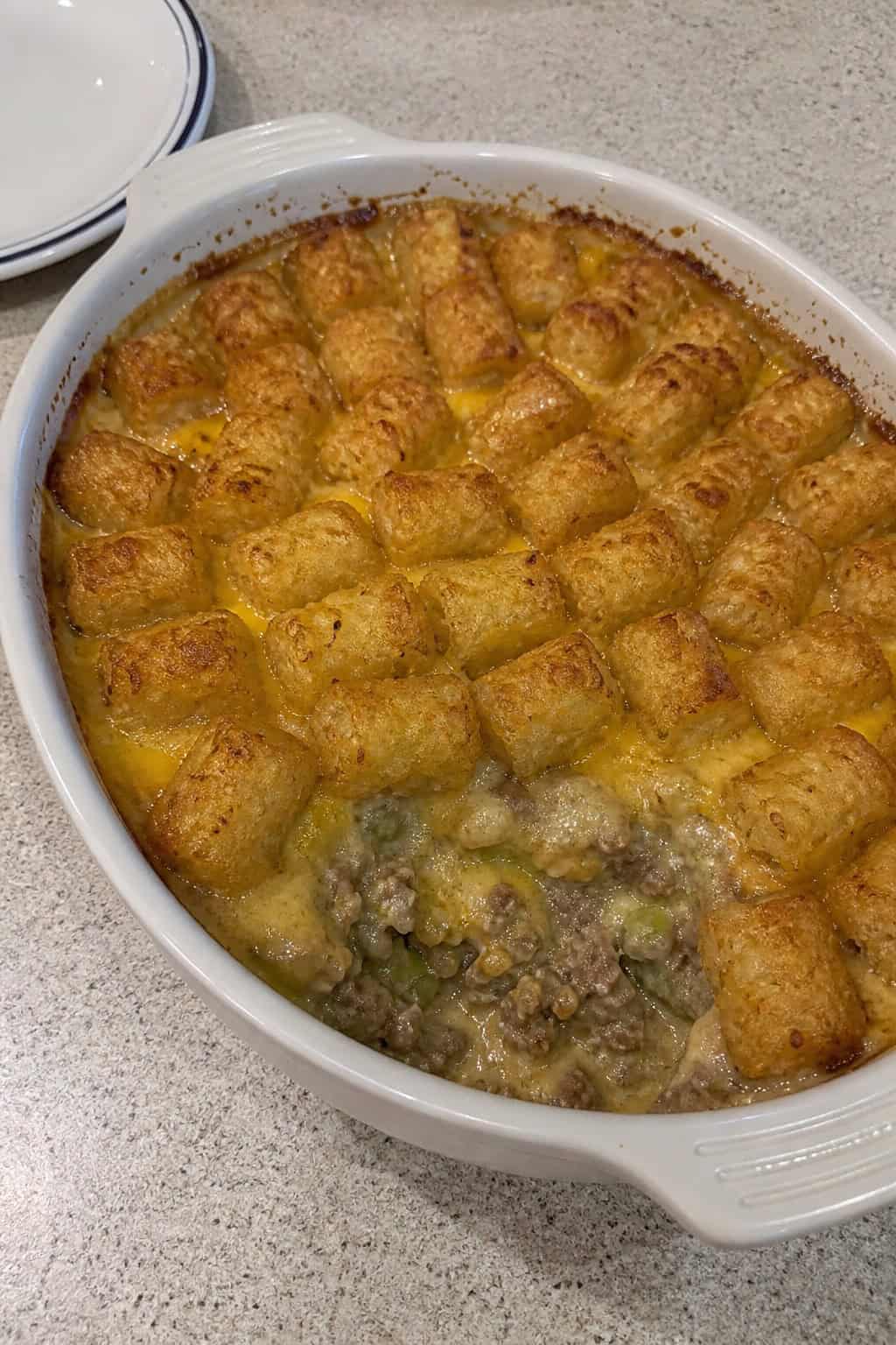 A freshly baked cheesy tater tot casserole in a worn rectangular baking dish on a wooden table, with a serving spoon resting in the corner and a small bowl of salad just visible at the edge.