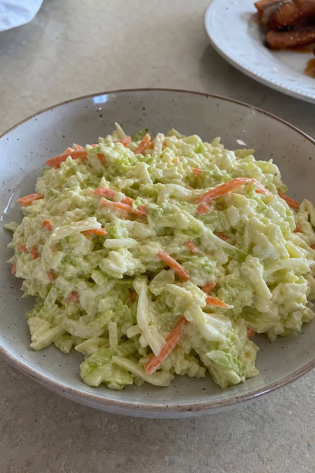 A bowl of creamy coleslaw with shredded cabbage and carrots on a wooden table, surrounded by fresh ingredients including cabbage leaves, carrot sticks, apple cider vinegar, and sugar.