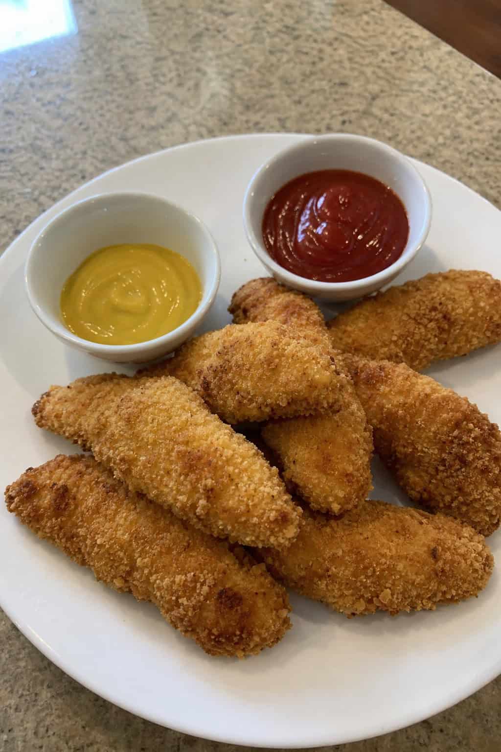 Golden panko-crusted chicken tenders arranged on a wooden board with two small dipping sauce bowls.