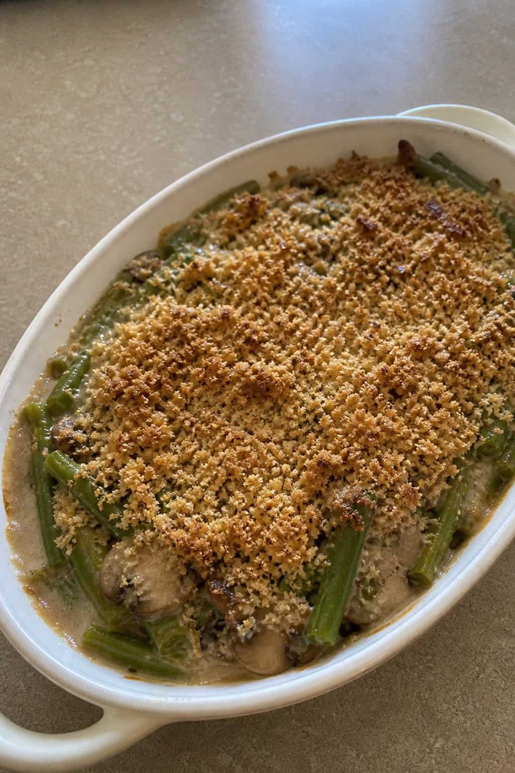 A green bean casserole with sliced mushrooms and a golden crispy onion topping in a white ceramic dish, surrounded by raw green beans and whole mushrooms on a wooden table.