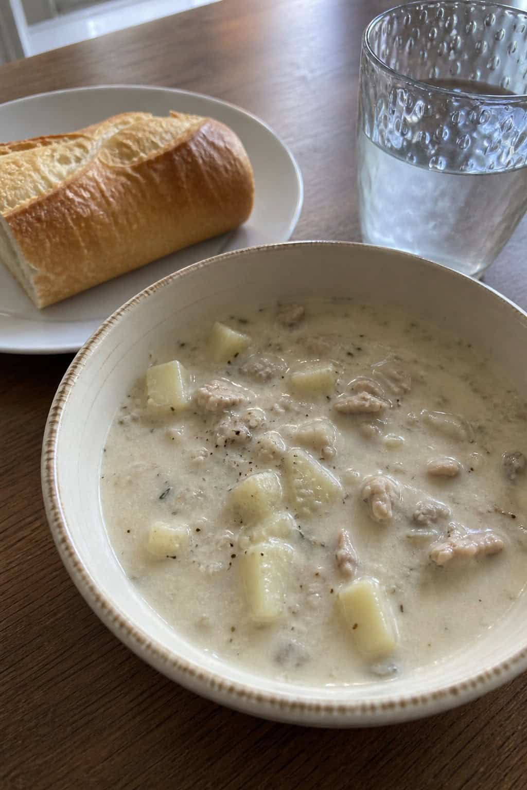 A steaming bowl of creamy New England clam chowder with clams and potatoes, surrounded by fresh ingredients and bread on a wooden table.