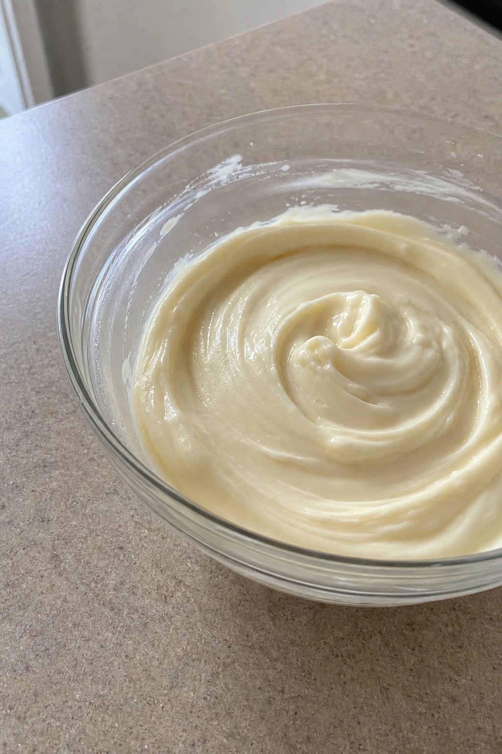 A glass bowl filled with smooth creamy cheesecake filling surrounded by cream cheese, eggs, sugar, and a vanilla bean on a wooden countertop.