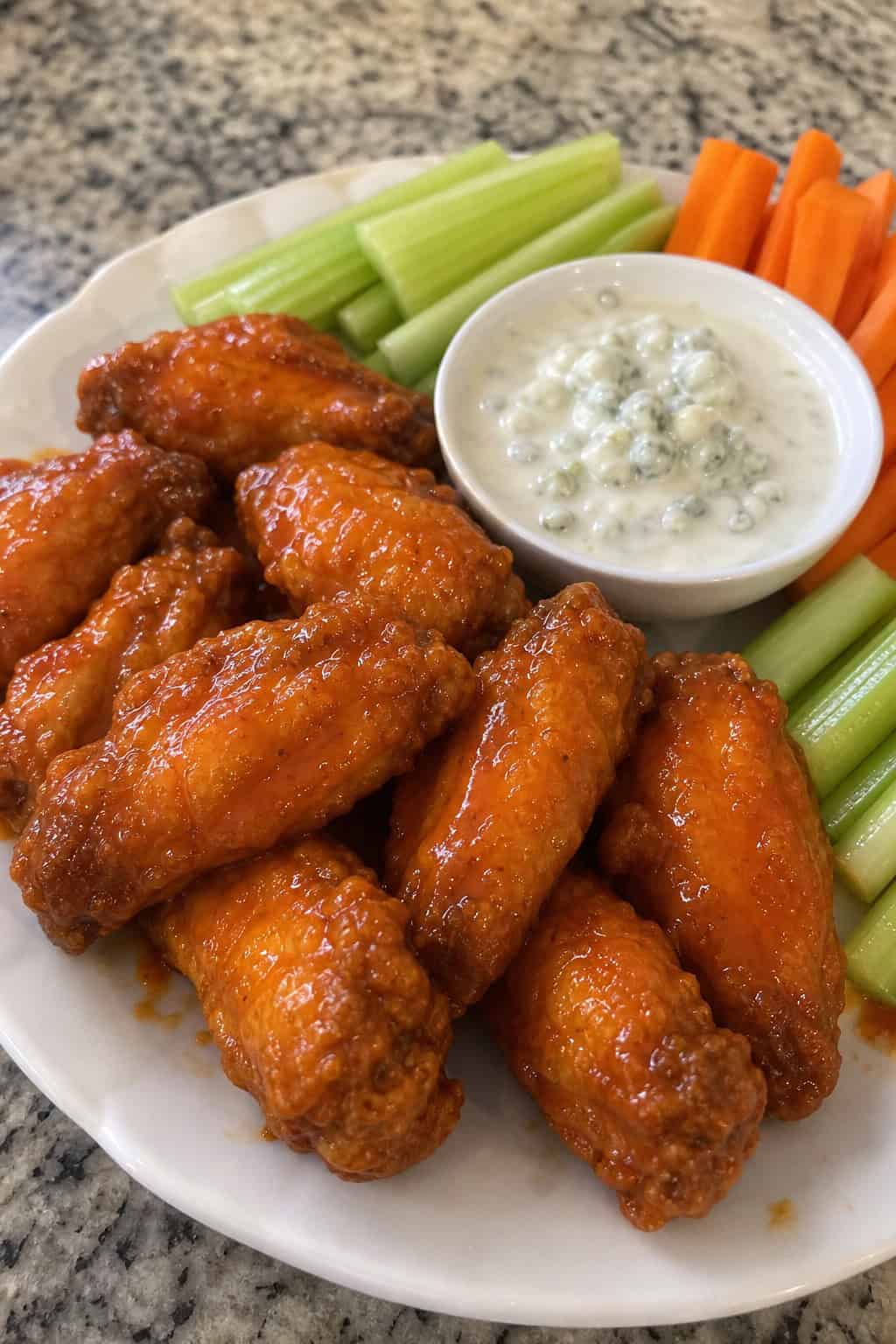 A plate of crispy buffalo chicken wings with a bowl of blue cheese dip, celery sticks, and carrot sticks on a wooden table.