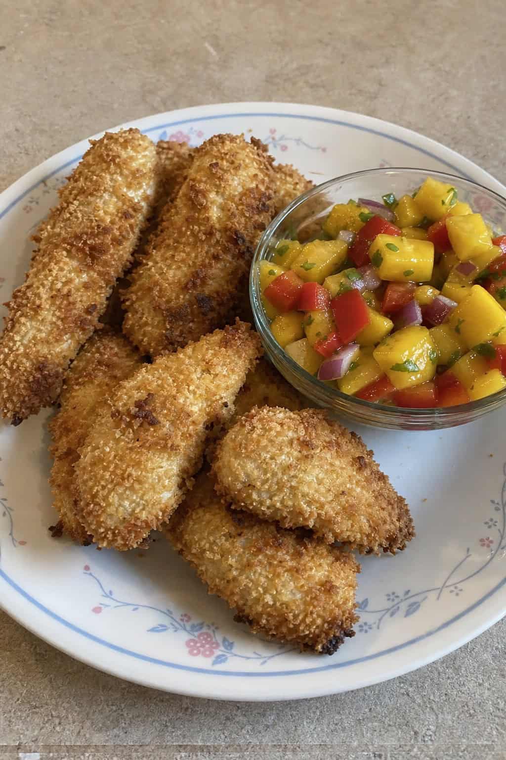 Plate of coconut-crusted chicken tenders served with a side of mango salsa and lime wedges on a wooden table.