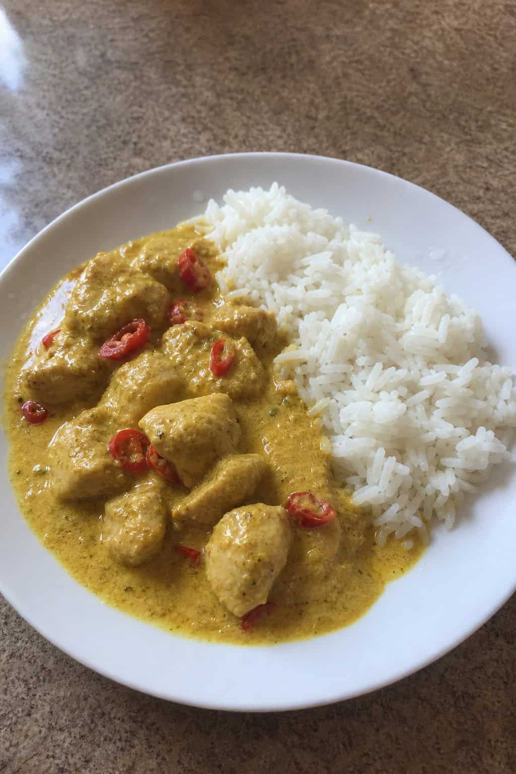 A plate of coconut curry chicken with jasmine rice garnished with cilantro and red chili on a wooden table.