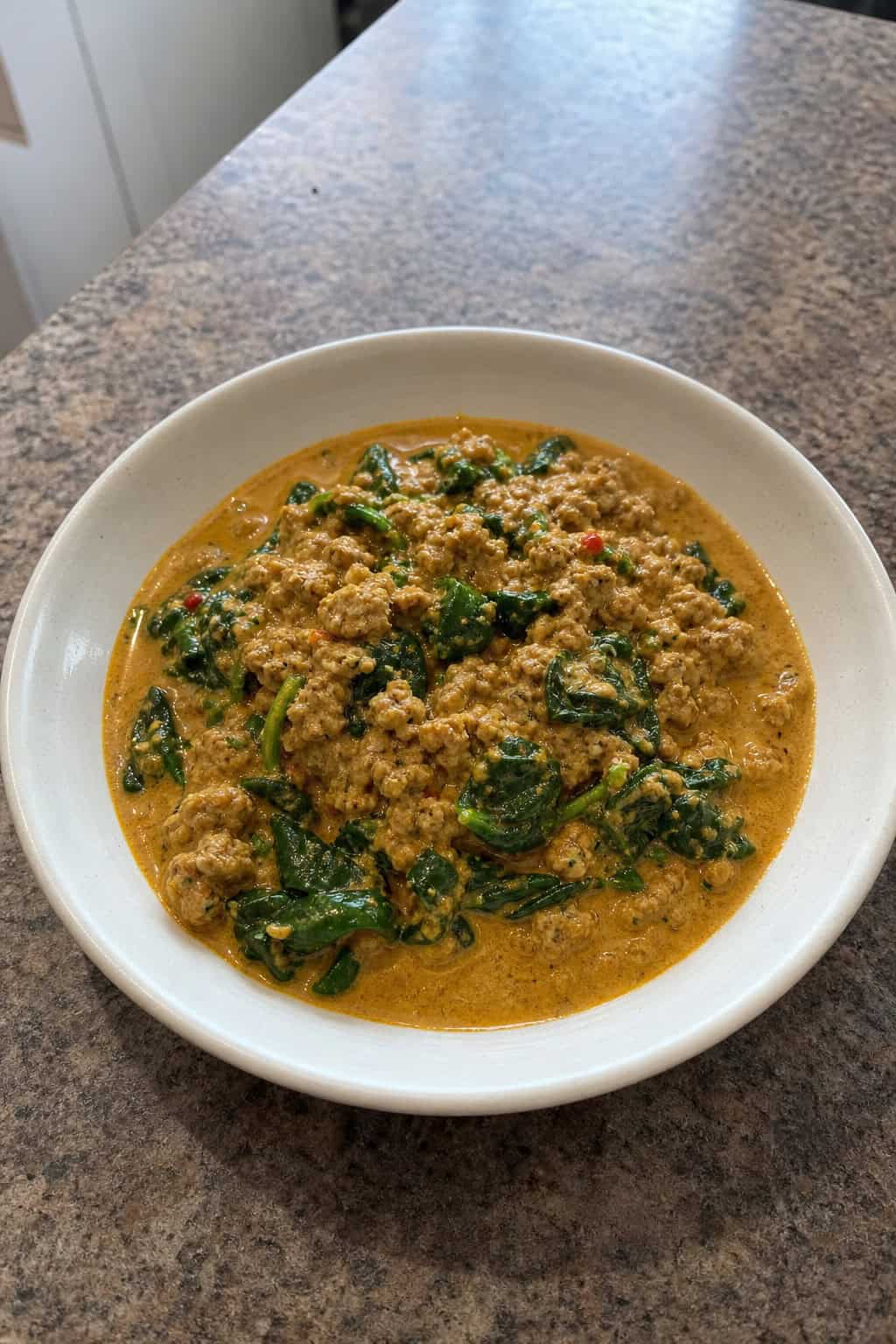 A bowl of coconut curry minced chicken with spinach on a wooden table, garnished with fresh herbs.