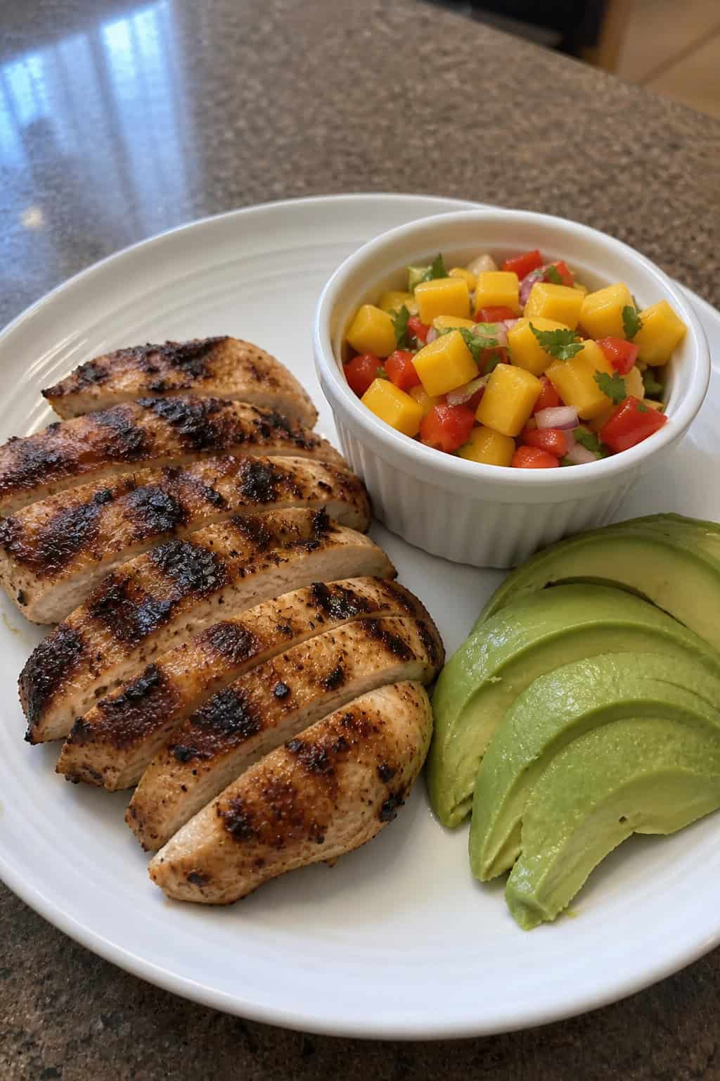 A plate with grilled chicken, mango salsa, and sliced avocado on a wooden table.