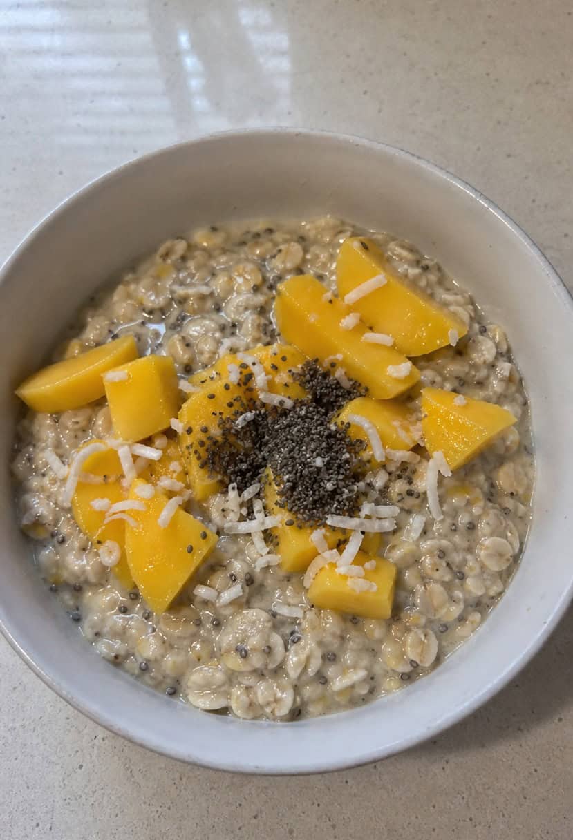 A bowl of coconut mango overnight oats surrounded by fresh mango slices, coconut shavings, and a sprinkle of chia seeds on a wooden table