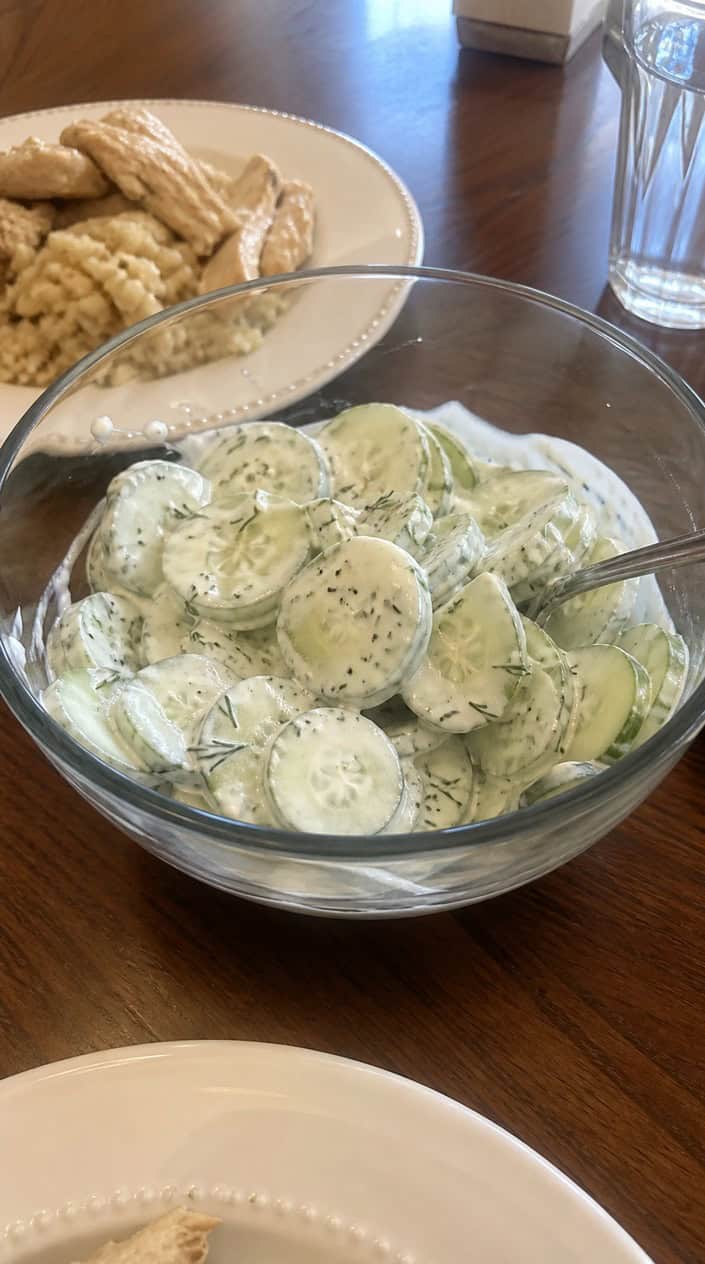 A bowl of cucumber-dill yogurt salad on a wooden table with fresh cucumbers and dill nearby.