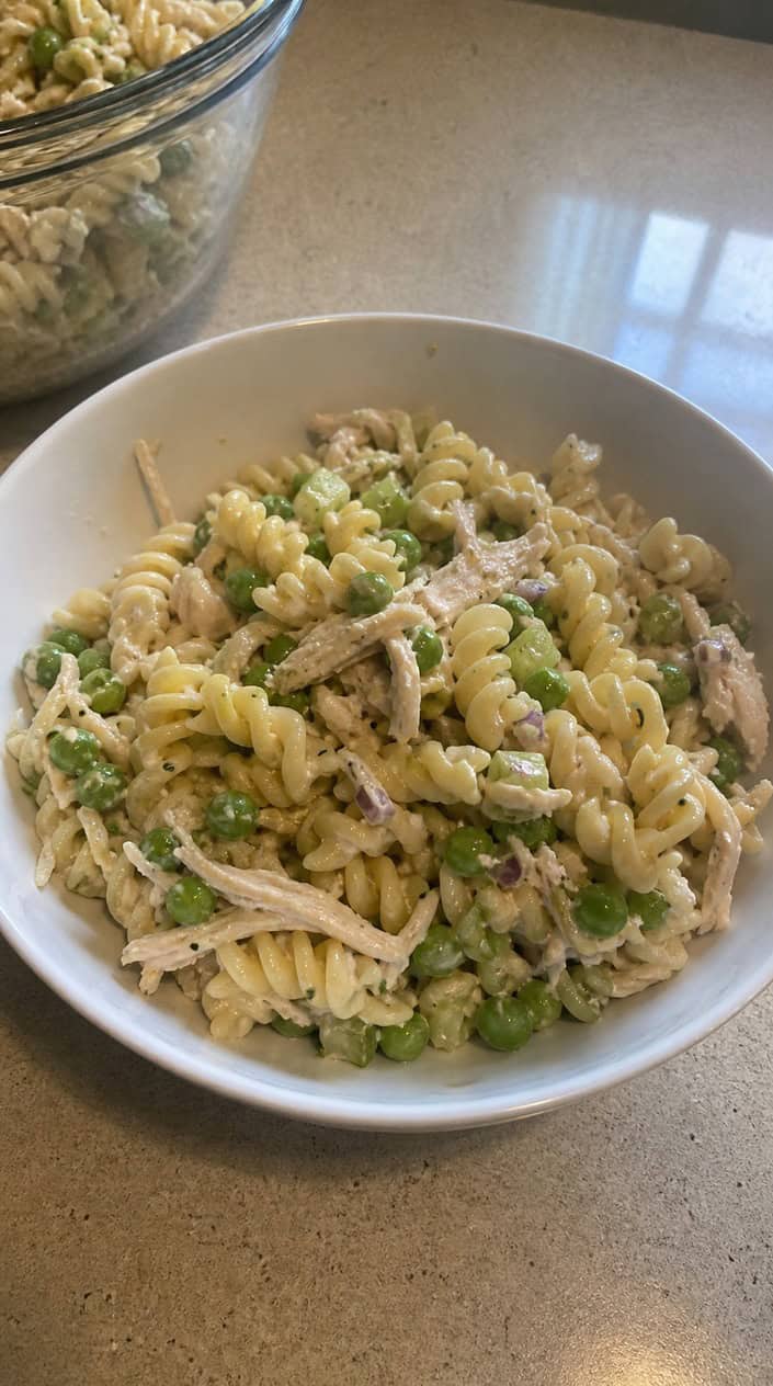 A bowl of cold rotisserie chicken pasta salad with peas on a wooden table, surrounded by lemon wedges and kitchen items.