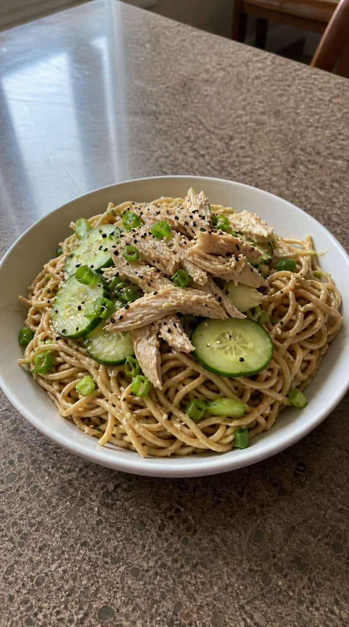 A bowl of cold sesame noodle salad with sliced cucumber and shredded rotisserie chicken on a wooden table with fresh ingredients around it.