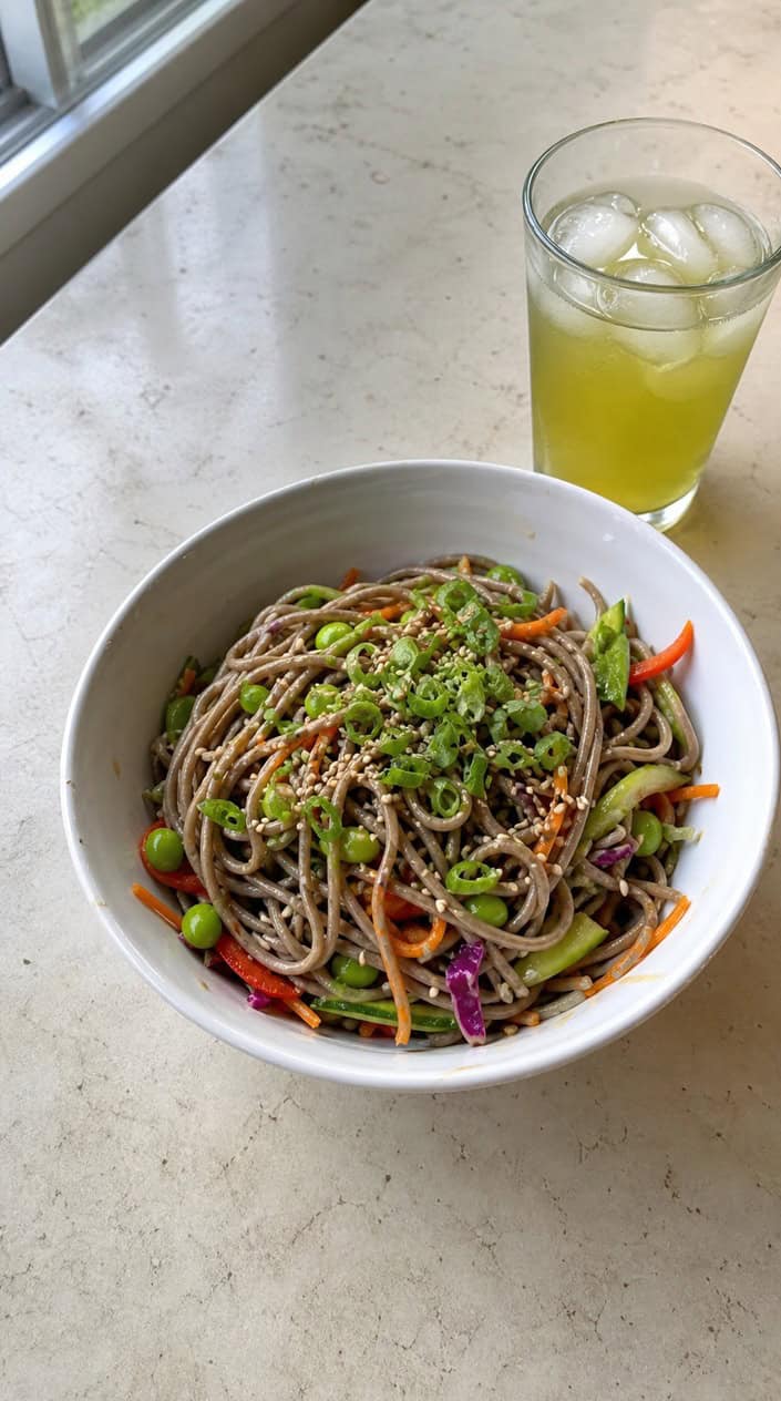 A bowl of cold sesame soba noodle salad garnished with sliced scallions on a wooden table with chopsticks and a glass of iced tea nearby.