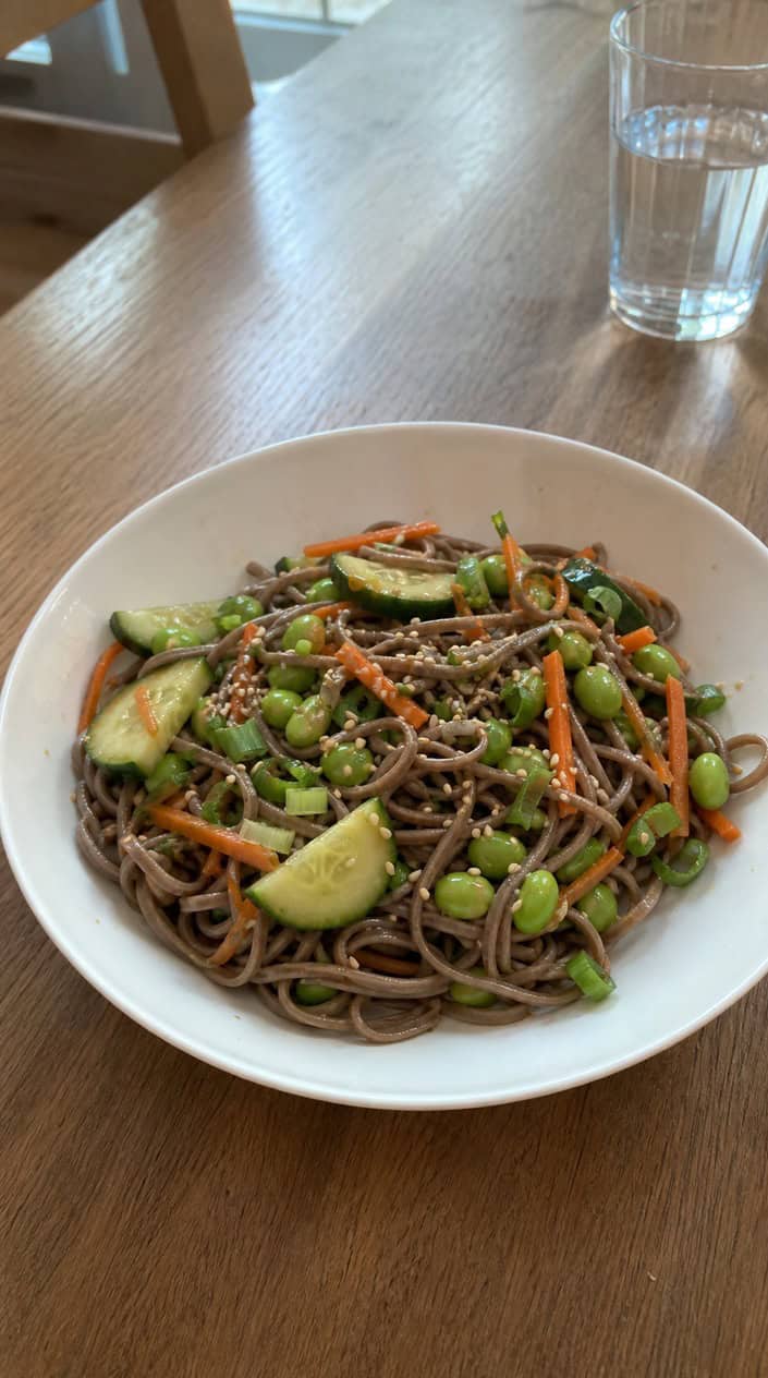 A bowl of cold soba noodle salad with sesame dressing, garnished with vegetables and sesame seeds, set on a wooden table outdoors with subtle Fourth of July decorations.