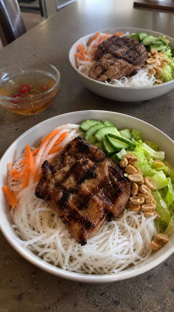 Two bowls of cold Vietnamese rice noodles with grilled pork chops, fresh herbs, and vegetables on a wooden table with dipping sauce and lime wedges.