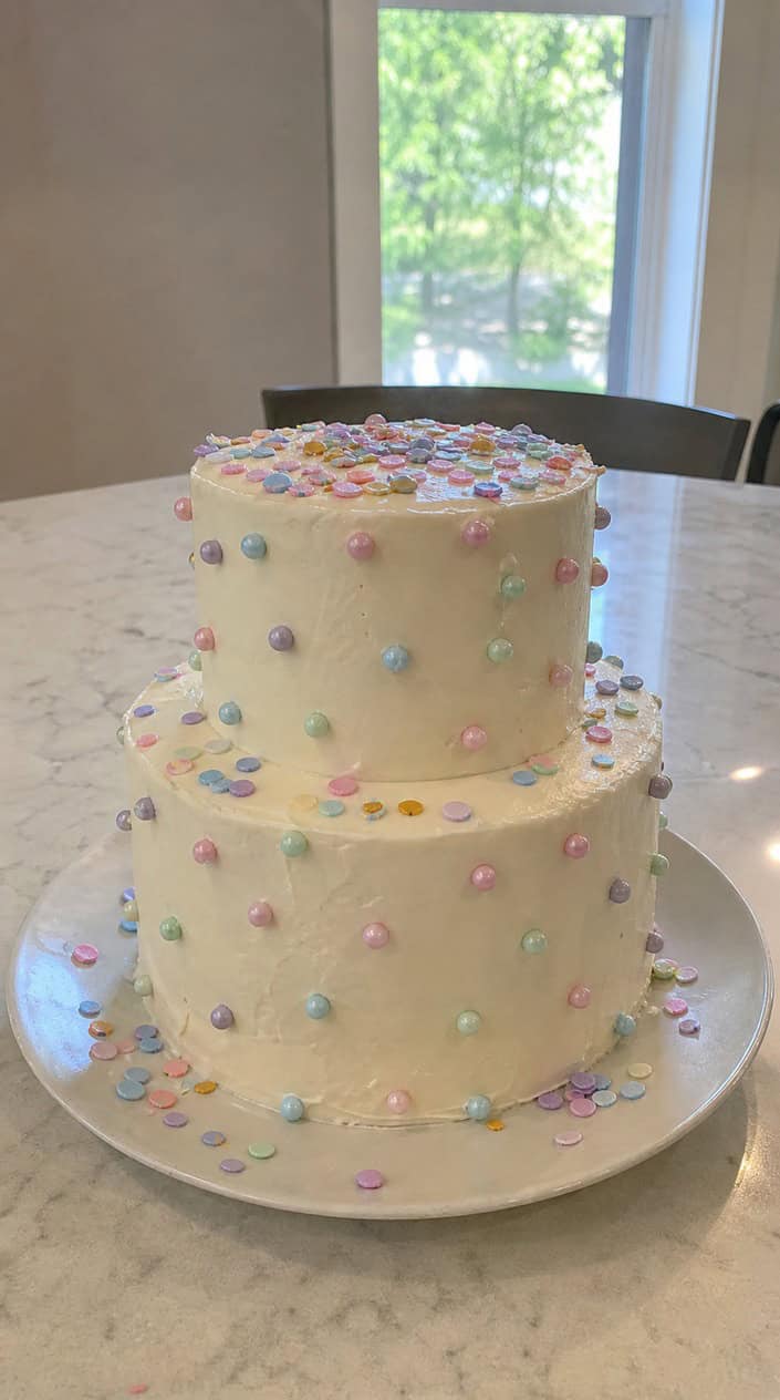 A gender reveal cake with white frosting, pastel candy pearls, and colorful confetti bursting from the center.