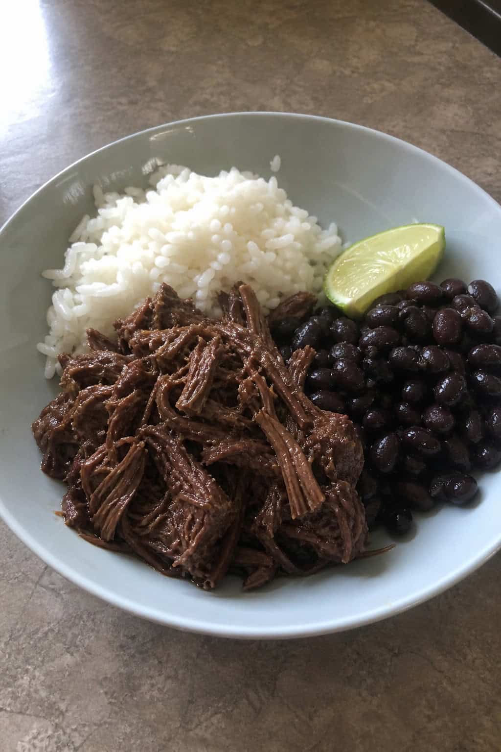A bowl of shredded chipotle barbacoa beef served with rice, black beans, and a lime wedge on a wooden table.