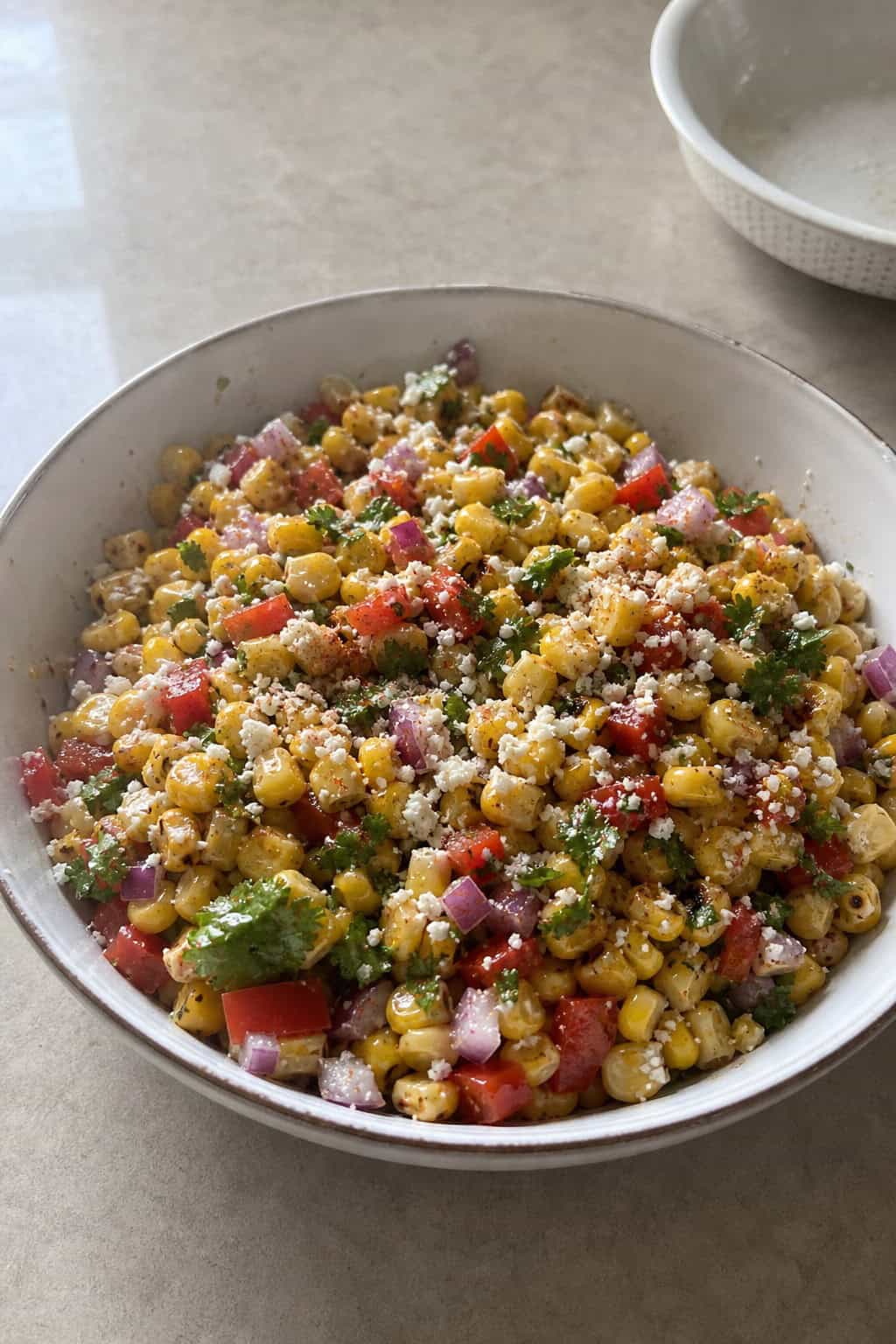 A bowl of colorful corn salsa with grilled corn, red onions, cilantro, red peppers, and cheese on a wooden table with lime wedges nearby.