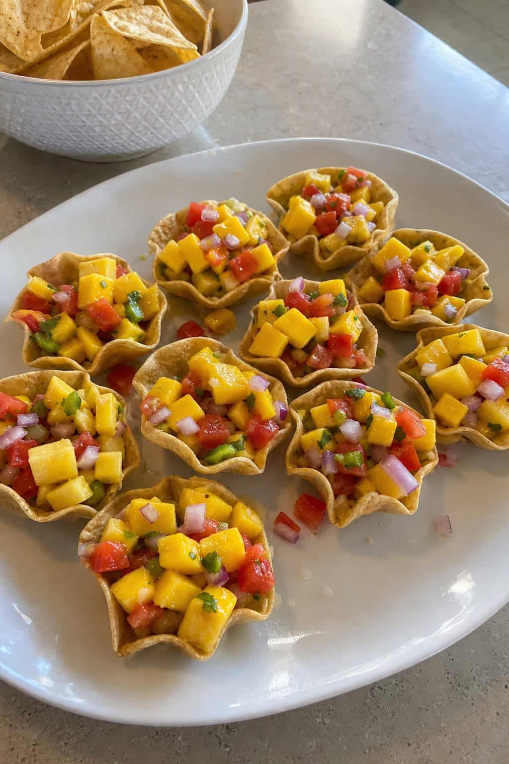 Clear tortilla chip cups filled with colorful mango salsa arranged on a wooden table with scattered tortilla chips and lime wedges.