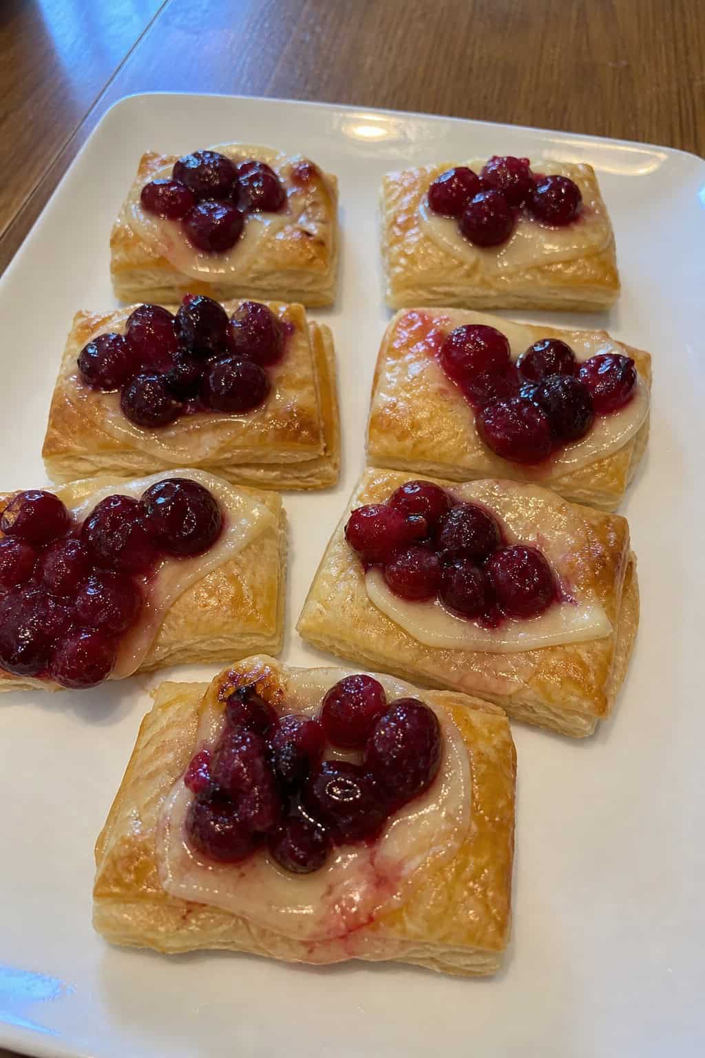 A platter of golden puff pastry bites topped with melted brie cheese and cranberries, garnished with herbs on a wooden table with holiday decorations in the background.
