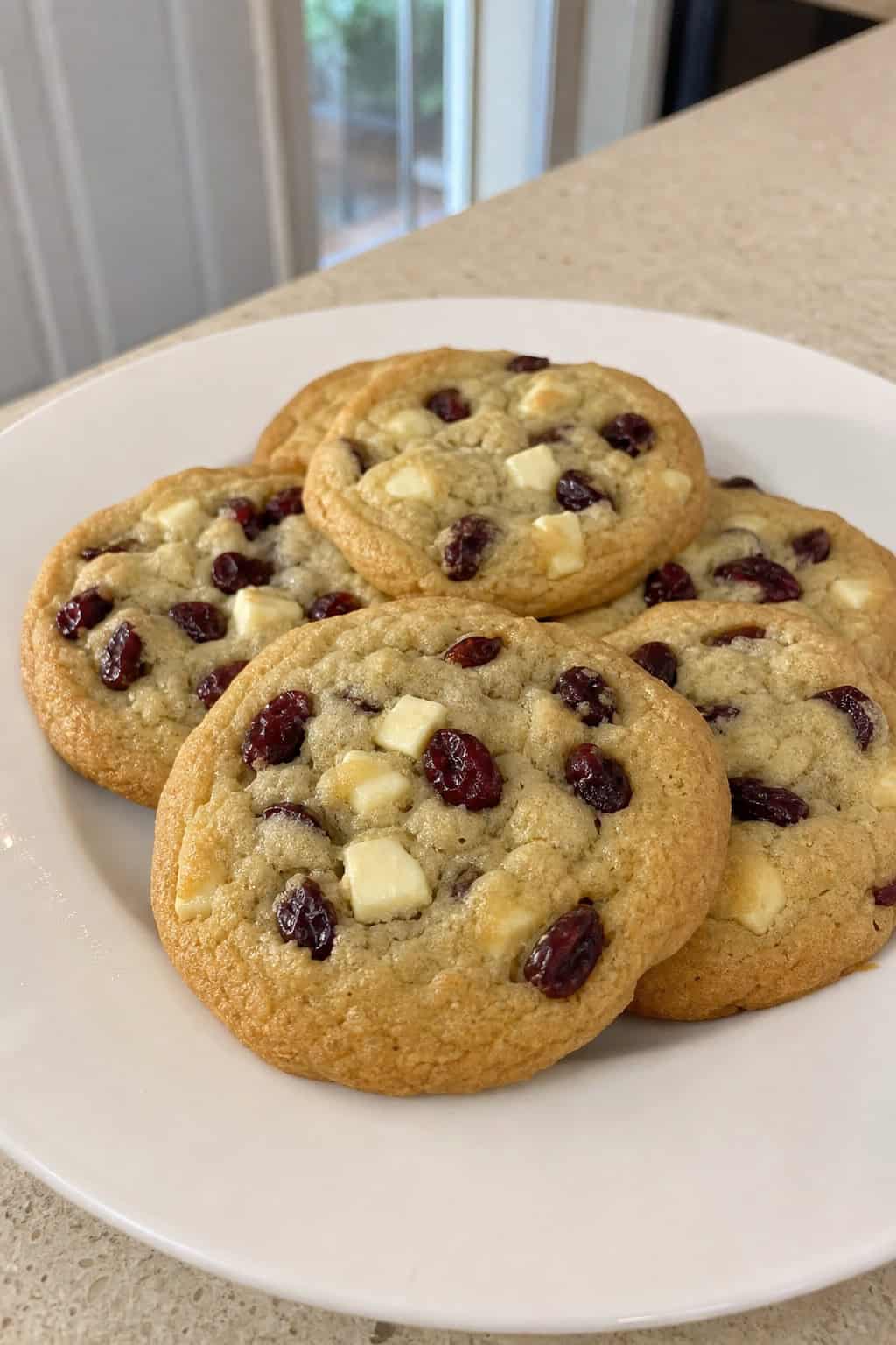 A close-up of cranberry white chocolate cookies on a wooden board with cranberries and white chocolate pieces around them.