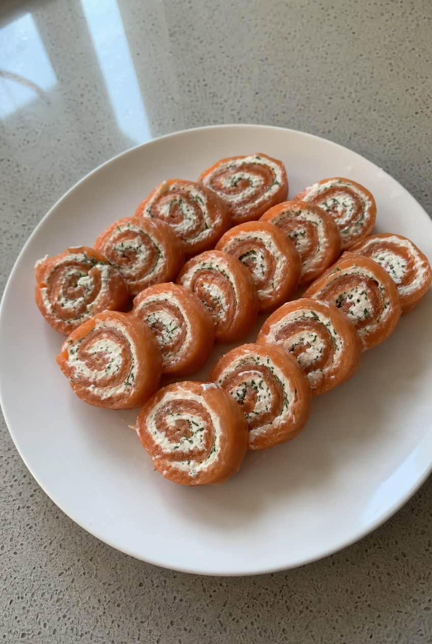 A wooden serving board with rows of spiral-cut tortilla pinwheels revealing layers of cream cheese, pink smoked salmon, and flecks of fresh dill, with lemon wedges and a small bowl of cream cheese alongside.