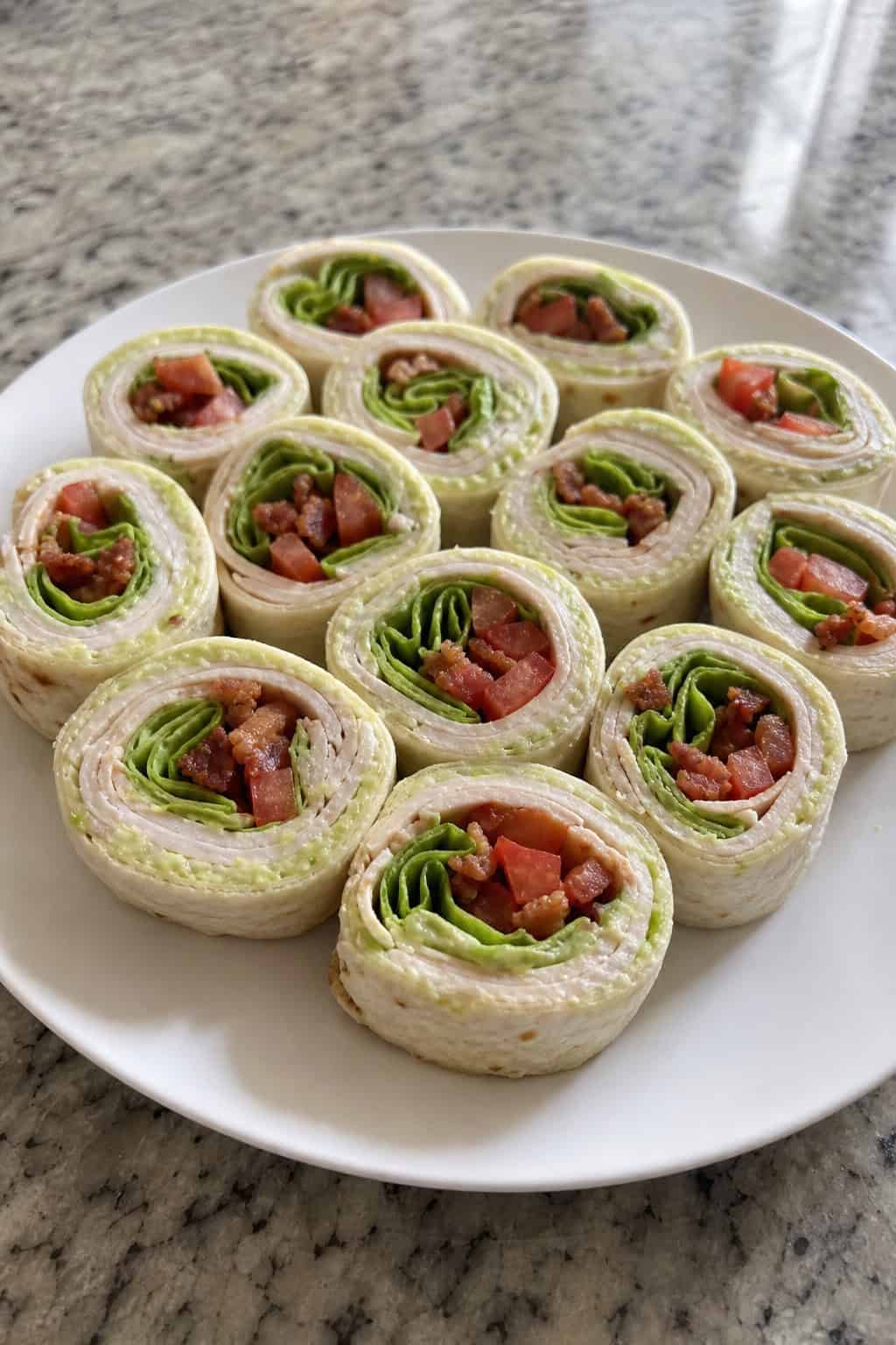 A plate of sliced creamy avocado turkey BLT pinwheel appetizers on a wooden board showing pale green avocado cream cheese filling with deli turkey, chopped bacon, and shredded lettuce layers, in a casual game day setting.