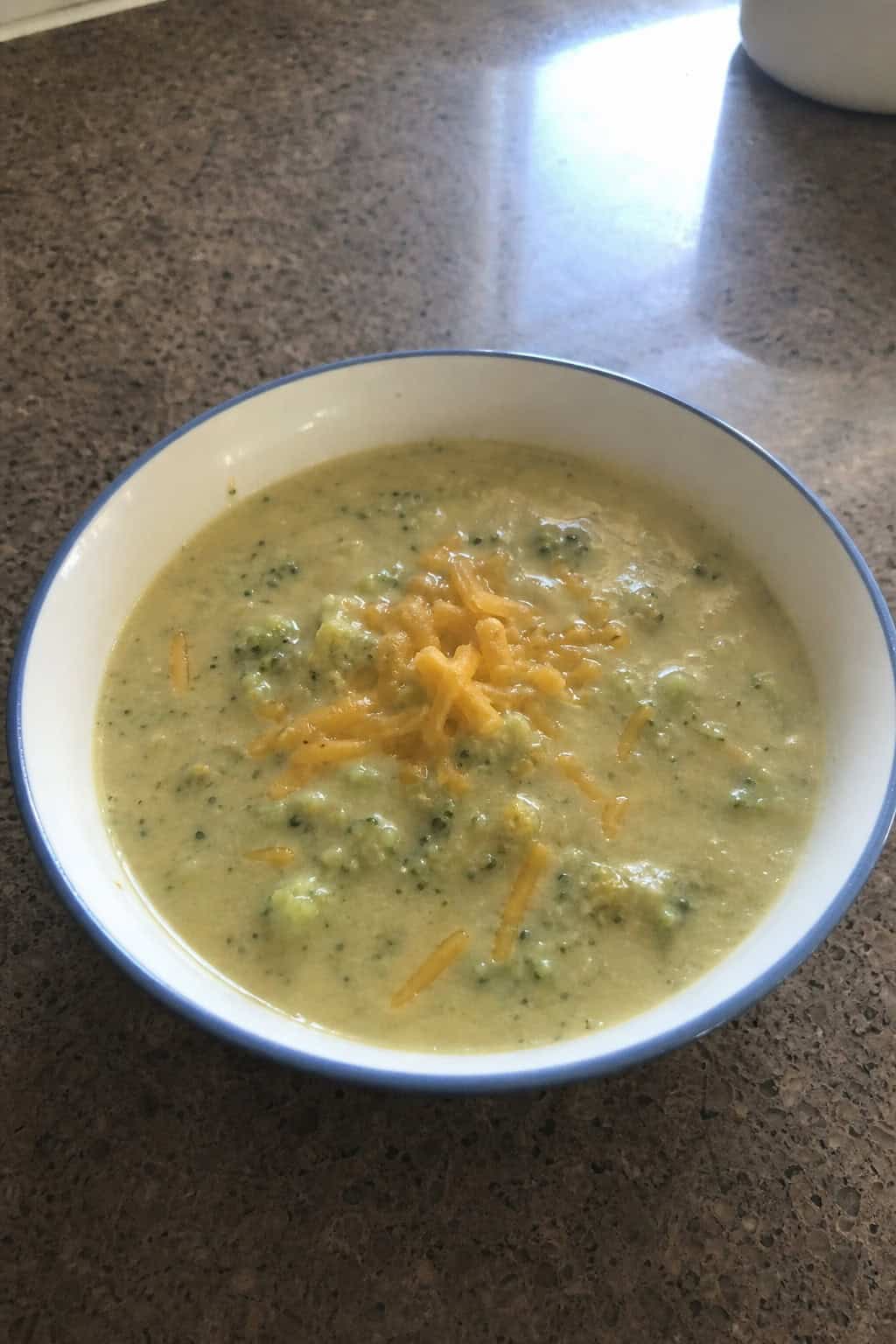 A bowl of creamy broccoli cheddar soup on a wooden table with fresh broccoli and shredded cheese nearby.