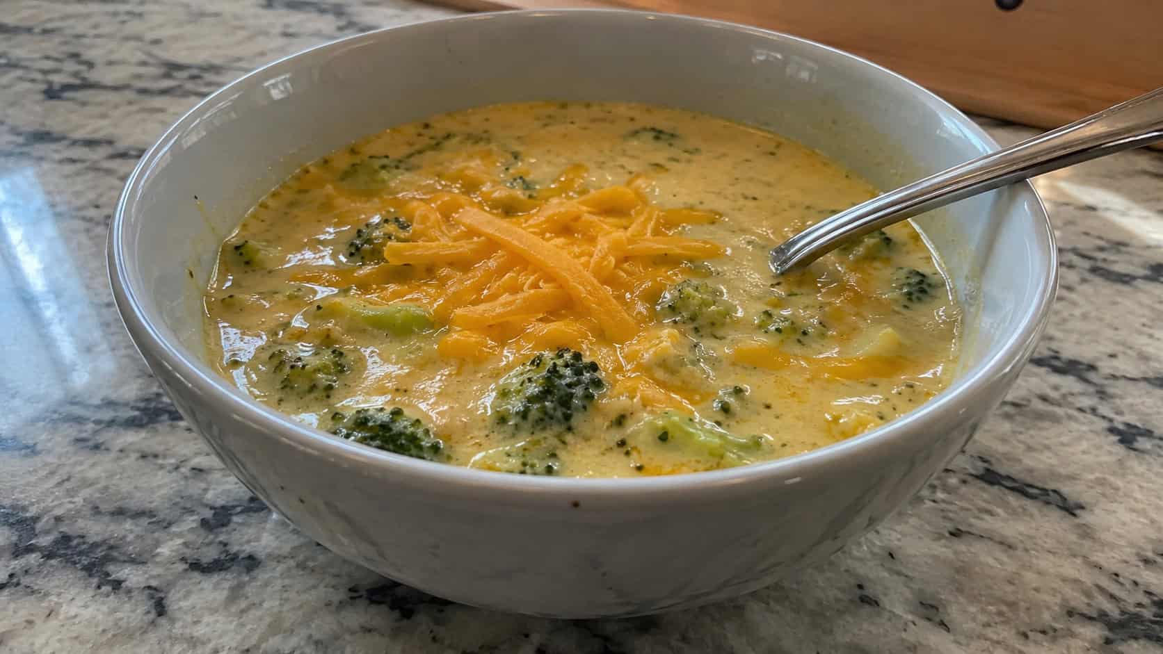 a finished bowl of creamy broccoli cheddar soup on a kitchen counter, visible tender broccoli florets, a spoon resting in the bowl, shredded sharp cheddar melting on top, warm natural window light, casual home cooking