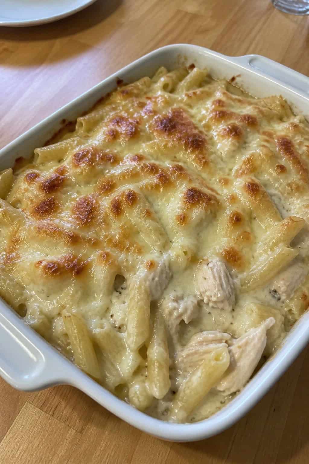 A creamy chicken Alfredo pasta bake in a white ceramic dish on a wooden table with fresh herbs and cheese around it.
