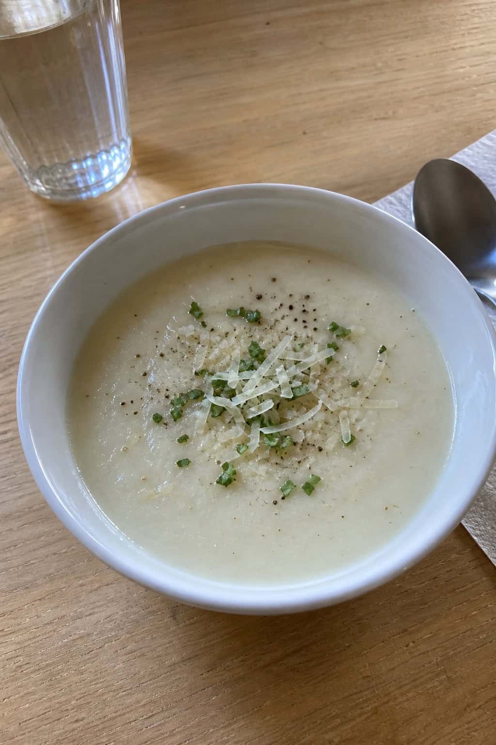 A bowl of creamy cauliflower and garlic soup garnished with Parmesan and parsley on a wooden table with fresh cauliflower and garlic nearby.