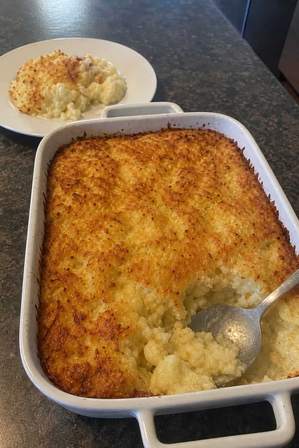 A creamy cauliflower and hashbrown casserole in a ceramic dish on a wooden table with a serving spoon.