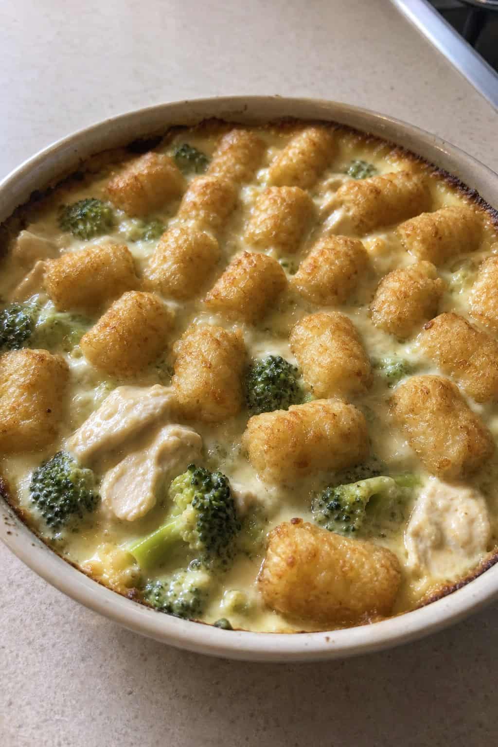 A creamy chicken and broccoli tater tot casserole in a deep rectangular baking dish, with golden tots covering the surface and a pale green filling visible at the edges.