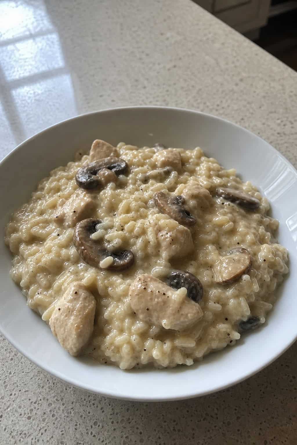 A wide shallow bowl of creamy risotto with shredded chicken and sliced cremini mushrooms folded throughout, finished with a sprinkle of fresh parsley, on a wooden table.
