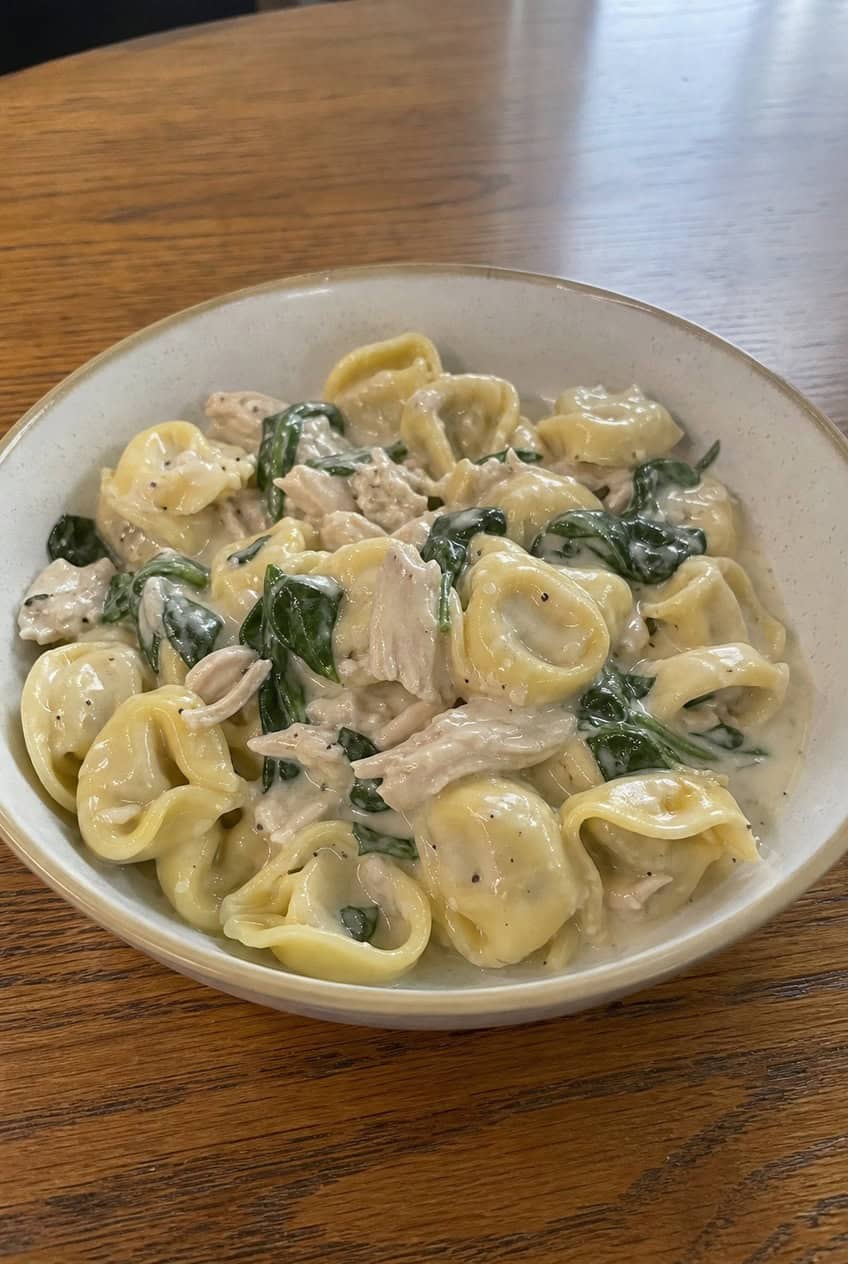 A shallow bowl of cheese tortellini in a creamy Parmesan sauce with wilted baby spinach and chunks of shredded rotisserie chicken, garnished with a few fresh spinach leaves, on a wooden table.