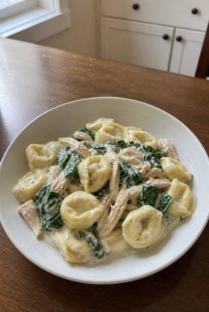 A shallow bowl of cheese tortellini in a creamy Parmesan sauce with wilted baby spinach and chunks of shredded rotisserie chicken, garnished with a few fresh spinach leaves, on a wooden table.