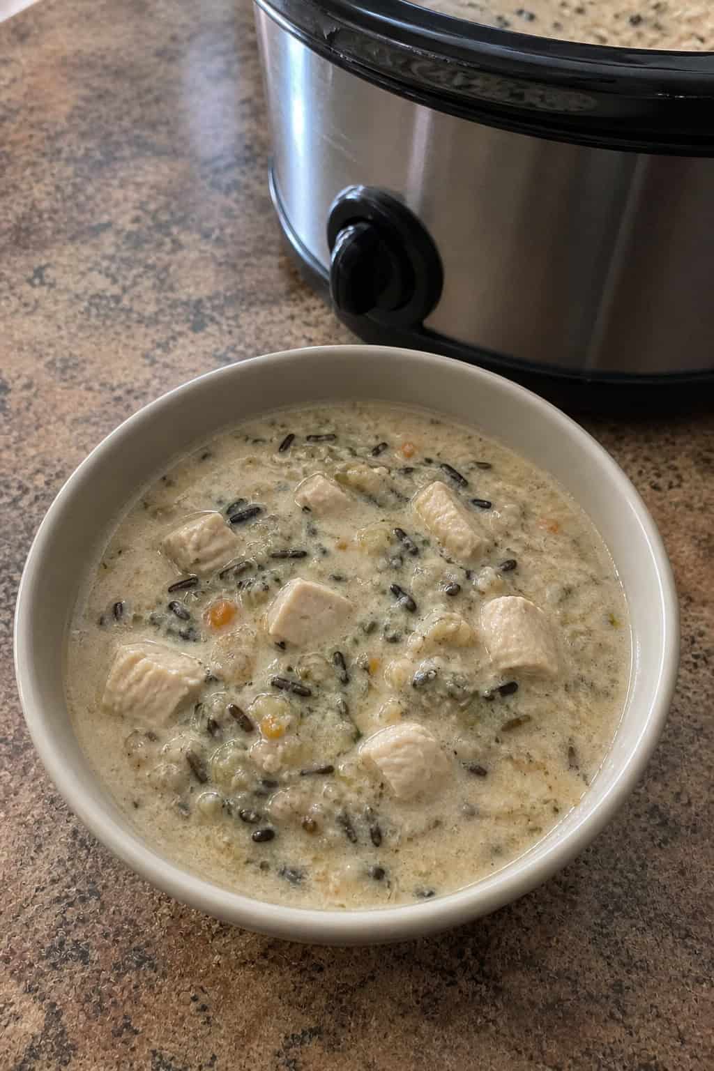 A bowl of creamy chicken and wild rice soup on a wooden table with fresh ingredients and a spoon nearby.